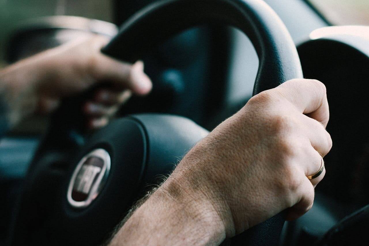 Close-up das mãos de uma pessoa segurando o volante de um carro. Uma mão está firmemente posicionada, enquanto a outra está levemente relaxada. O interior do carro é visível ao fundo, com detalhes desfocados.