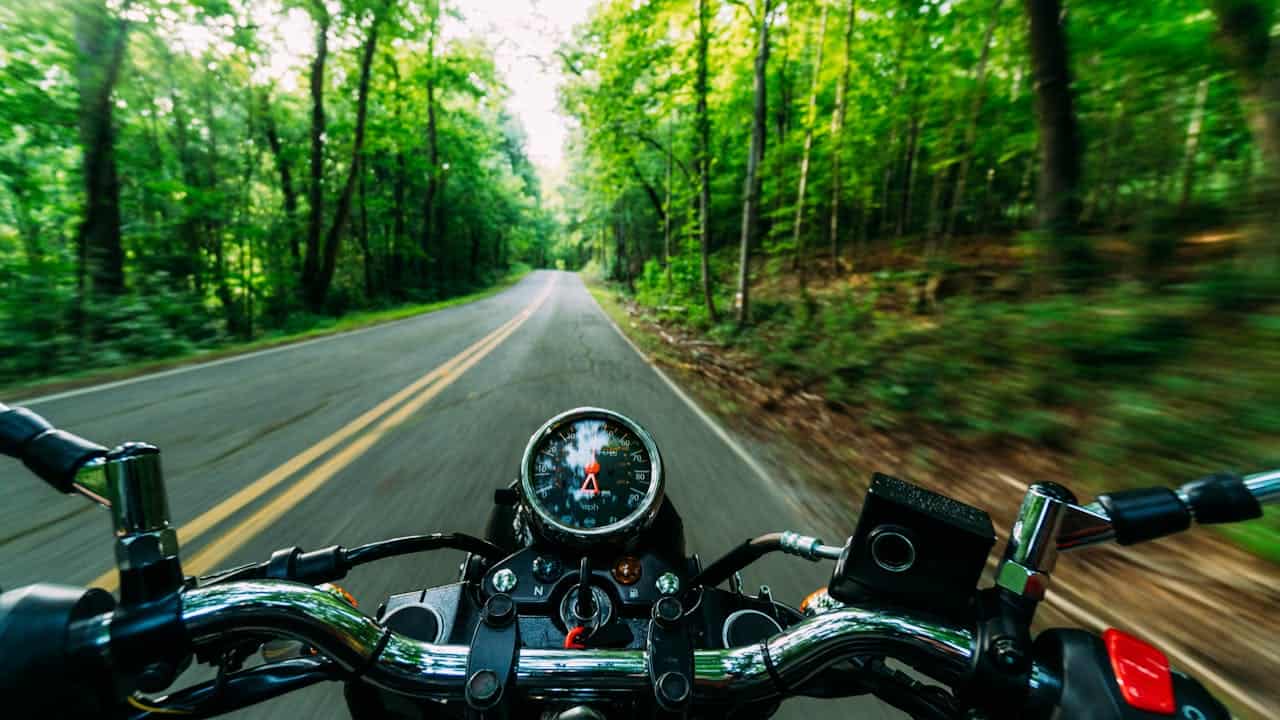 Pessoa pilotando uma moto por estrada arborizada, vista em primeira pessoa com velocímetro em destaque. 