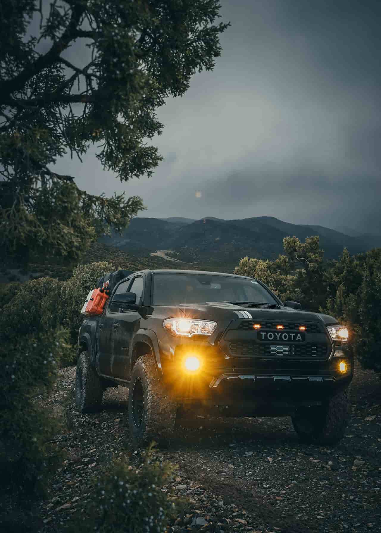 Picape Toyota 4x4 com faróis acesos em área montanhosa e vegetação densa, pronta para condução off-road.