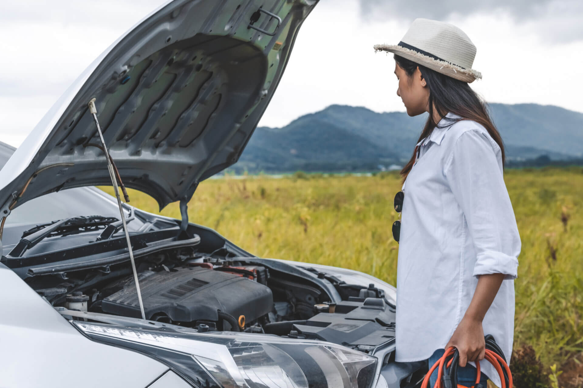 Mulher de roupa clara em frente a um carro de capô aberto. Nas mãos ela segura um cabo para faze chupeta elétrica.