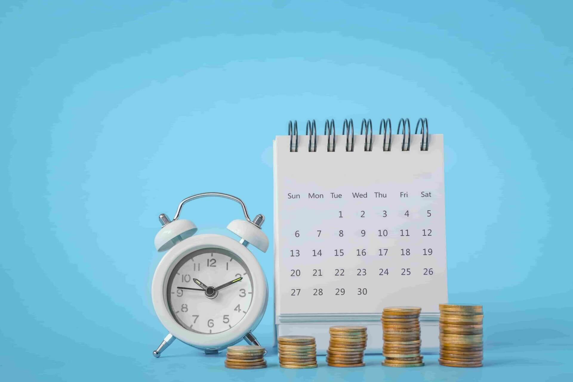 Relógio posicionado ao lado de um calendário e de pilhas de moedas douradas.