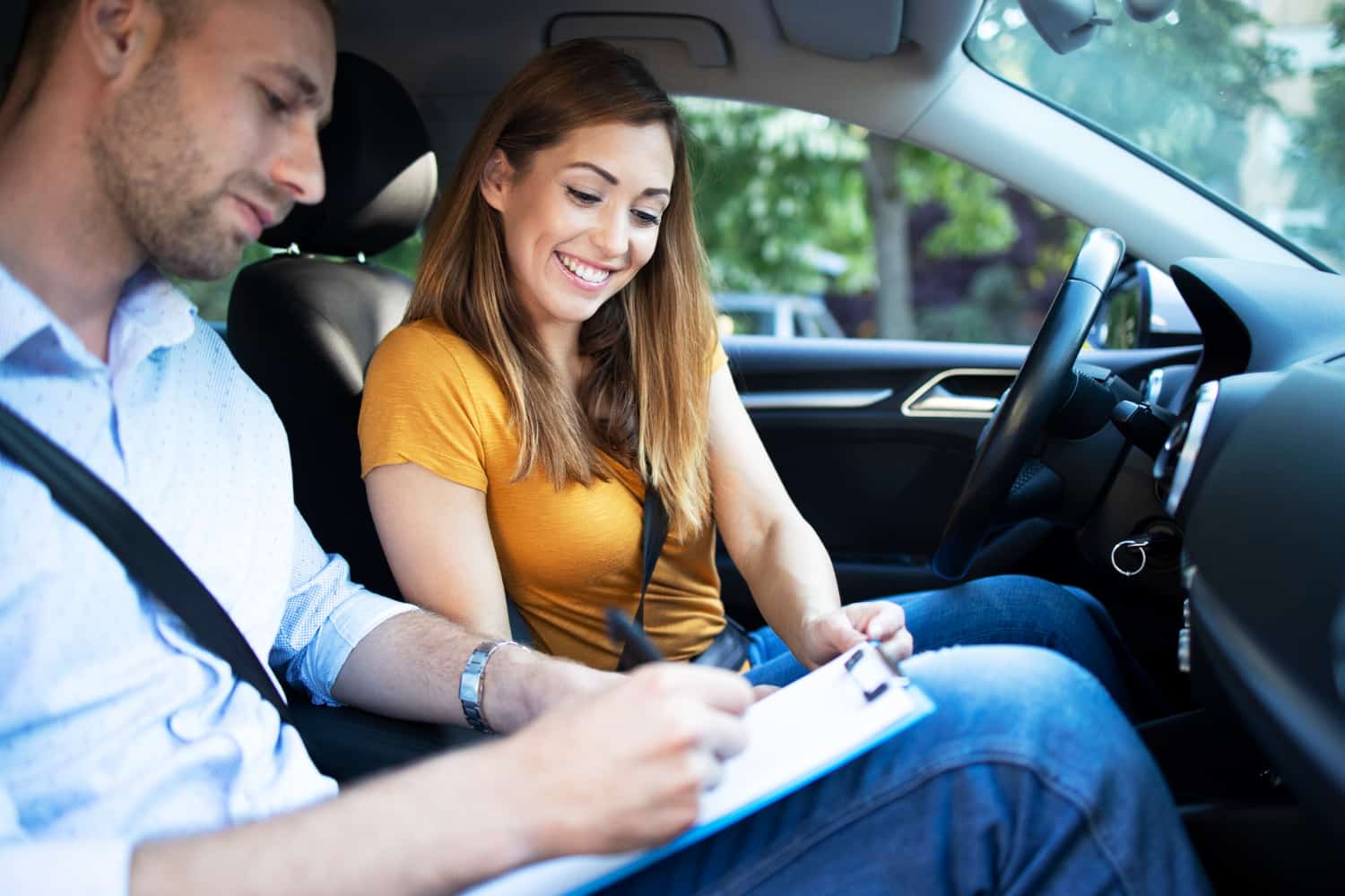 Homem e mulher sentados em um carro, com o homem segurando uma prancheta e uma caneta, enquanto a mulher sorri.