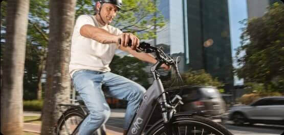 Homem pedalando uma bicicleta elétrica preta na rua da cidade, com arranha-céus ao fundo, durante o dia.