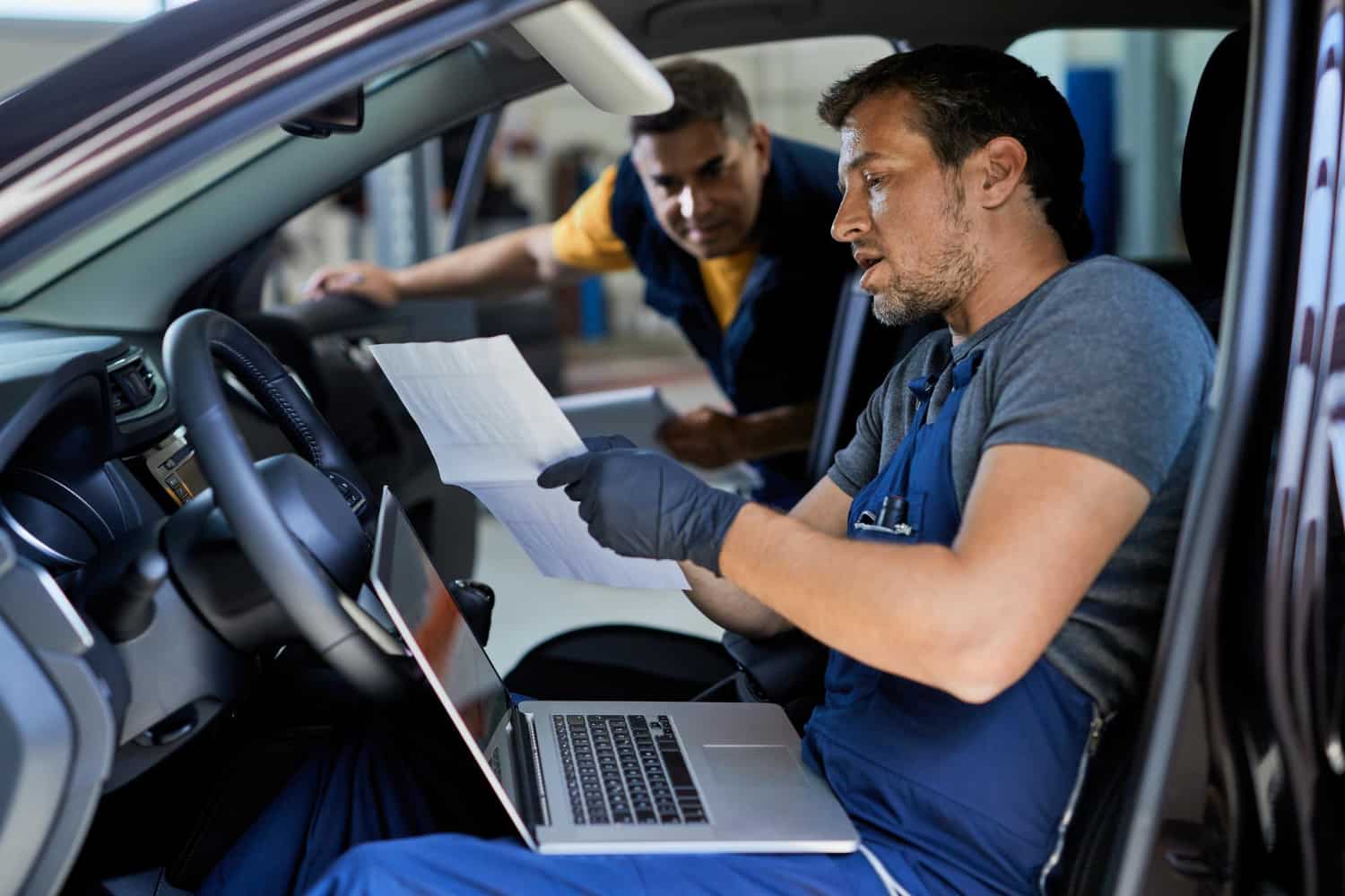 Dois homens em um carro, segurando uma folha de papel e uma caneta, em um ambiente de oficina de automóveis.