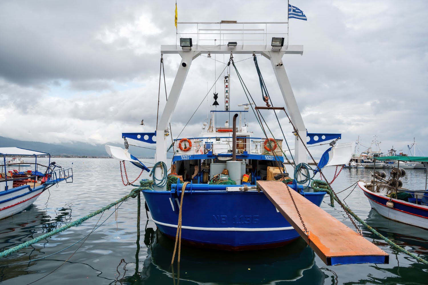 Barco de pesca de cor azul atracado na marina, com uma placa de acesso na frente, céu nublado e montanhas ao fundo.