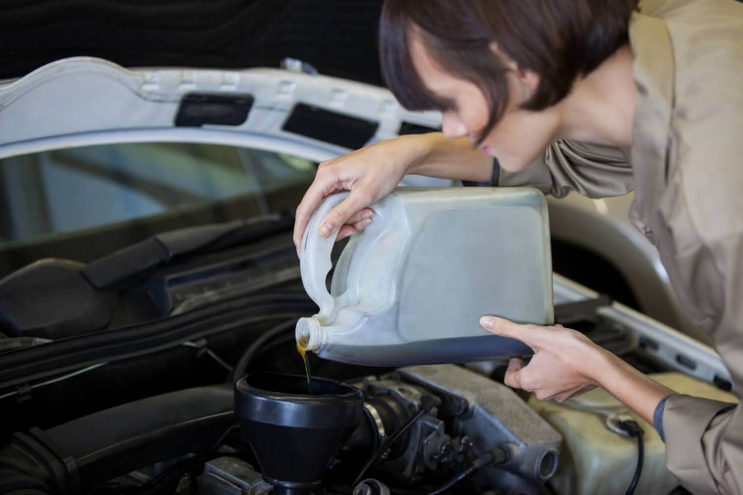 Pessoa enchendo o reservatório de óleo do motor de um carro, em um ambiente de oficina, usando um recipiente de plástico cinza.