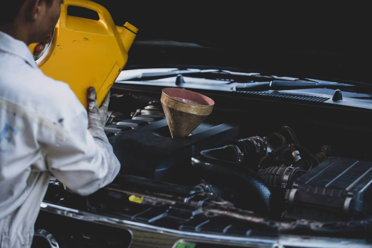 Pessoa despejando óleo automotivo de um recipiente amarelo enquanto faz manutenção no motor de um carro, em um ambiente de oficina.