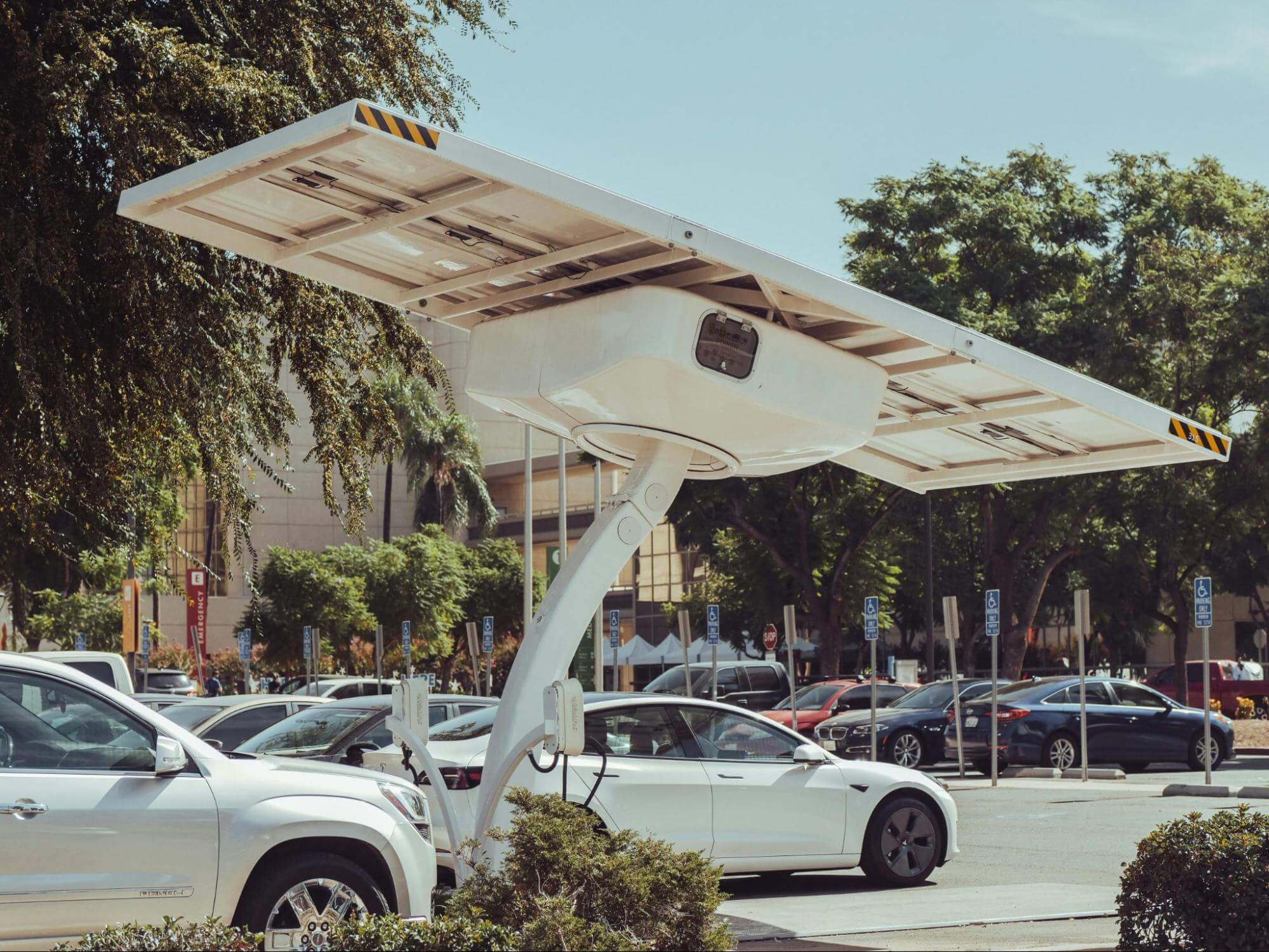 Estacionamento com diversos veículos e uma placa de energia solar com carregador para carros elétricos e híbridos. 