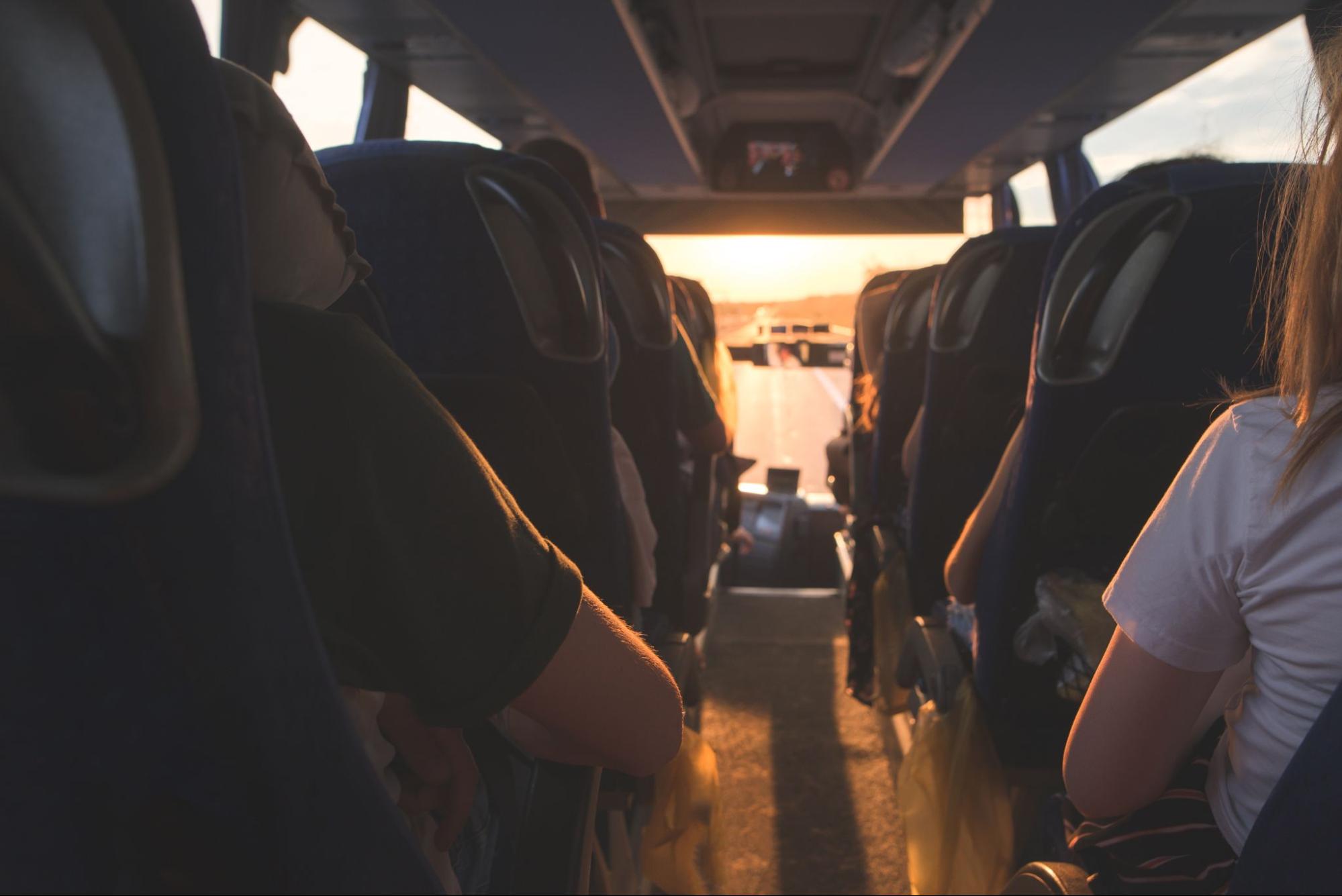 Pessoas sentadas em assentos de ônibus de costas. Ao fundo, o vidro do veículo mostra a estrada ao pôr-do-sol