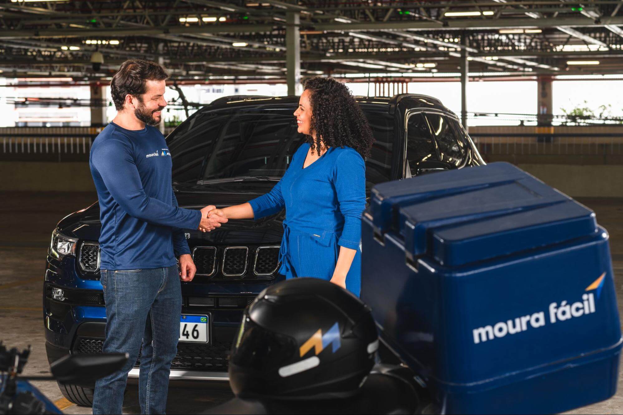 Homem com uniforme Moura aperta a mãos de uma mulher em um estacionamento. A moto com o logo Moura Fácil na frente dos dois e um carro escuro está atrás.
