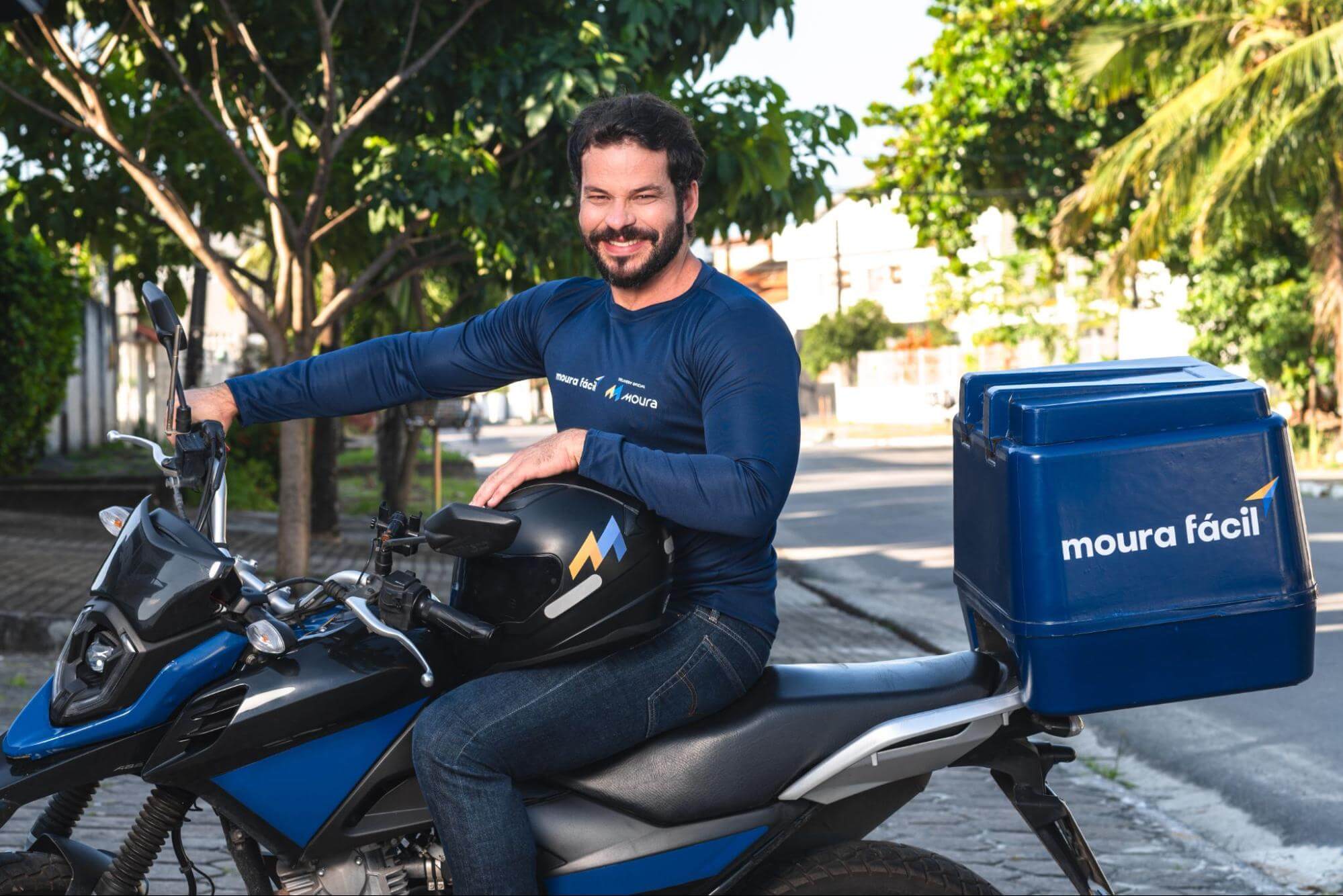 Homem sorridente em moto do Moura Fácil segura capacete no colo.