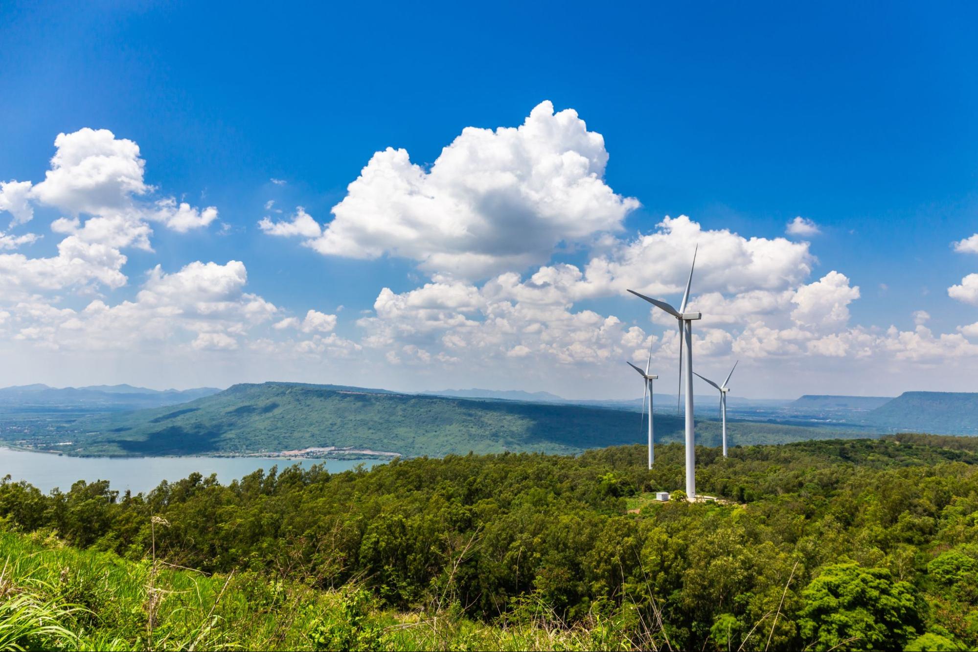 Três turbinas eólicas em uma colina verde com vista para um lago e montanhas, sob um céu azul com nuvens.