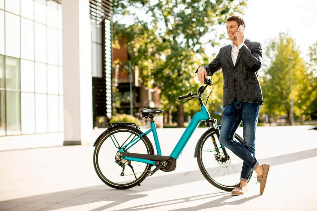 Homem de negócios falando ao telefone com o braço escorado em uma bicicleta elétrica.