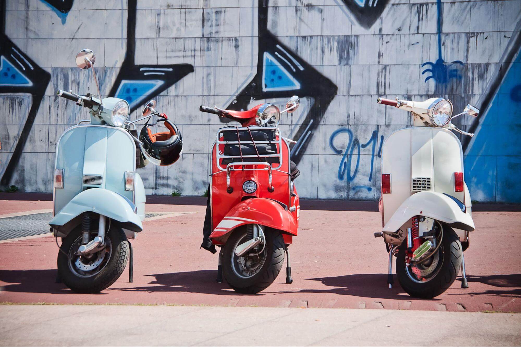 Três scooters de cores diferentes estacionadas uma ao lado da outra com muro grafitado ao fundo