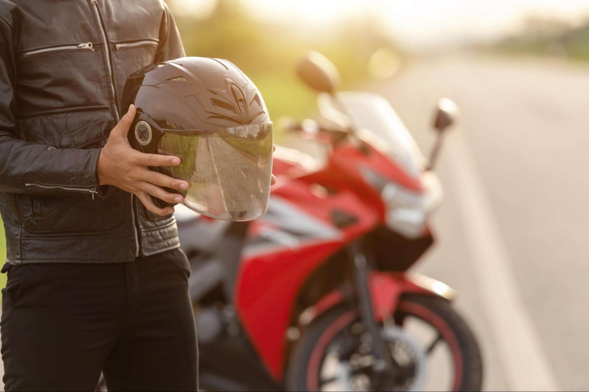Homem com jaqueta de couro segura capacete preto em suas mãos. Ao fundo, em desfoque uma moto vermelha no acostamento de uma estrada.