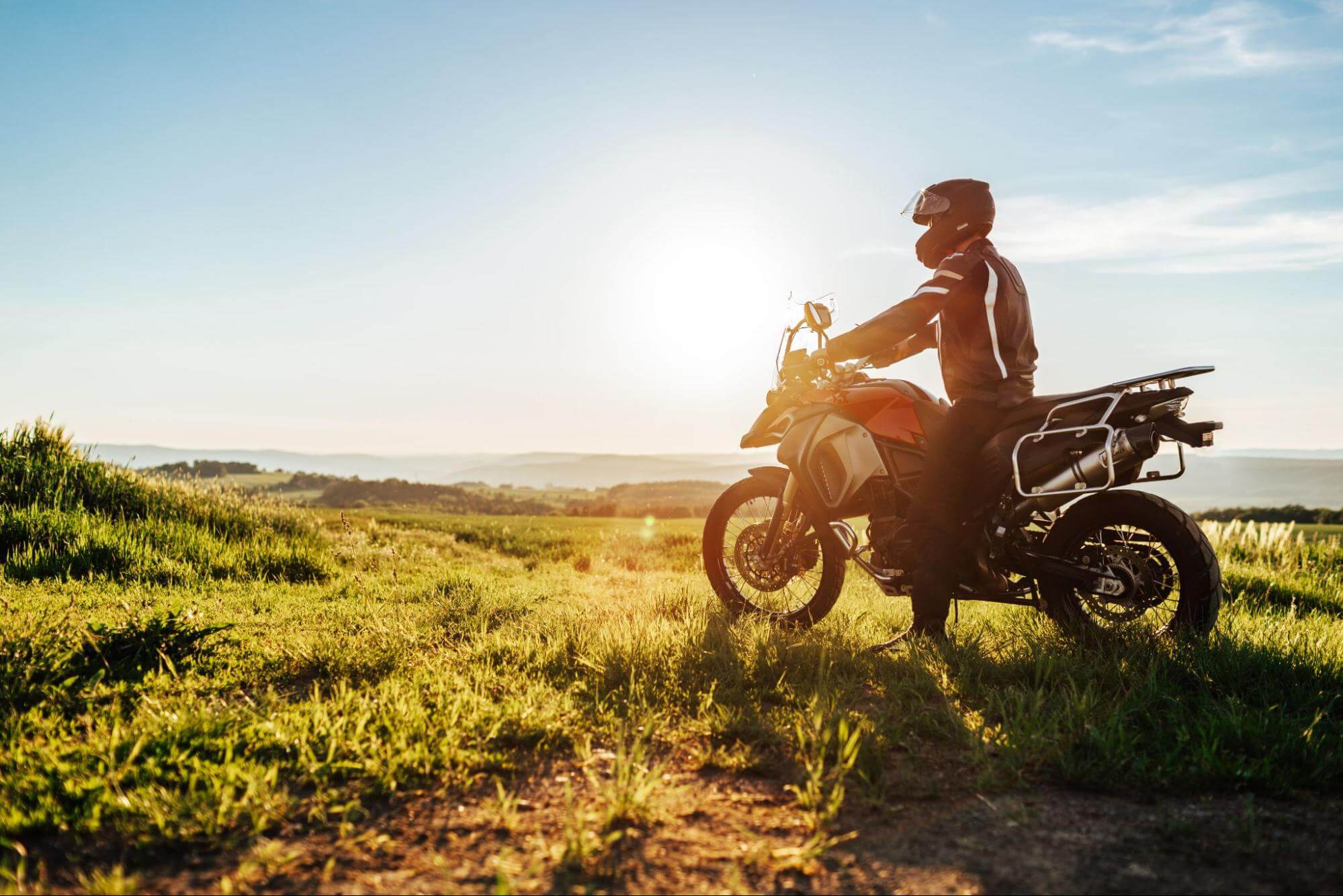 Motociclista de capacete e jaqueta montado em moto trail vermelha em campo verde, sob a luz do sol poente e céu claro ao fundo.