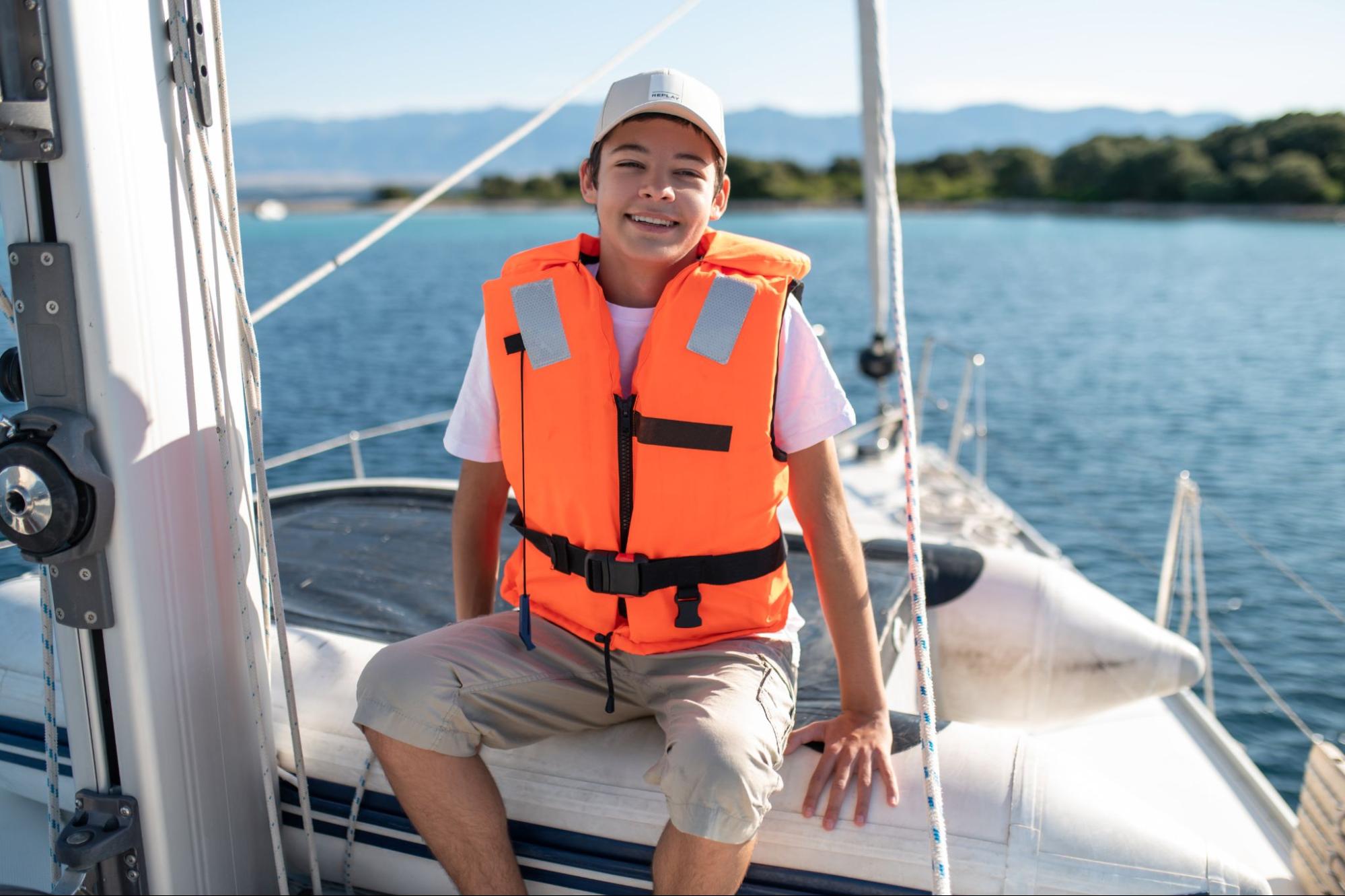 Menino sorridente sentado na ponta de um barco com colete salva-vidas laranja.