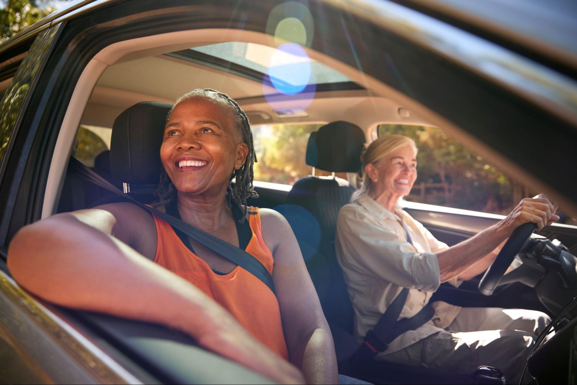 Duas mulheres mais velhas sorriem dentro de um carro em dia ensolarado.