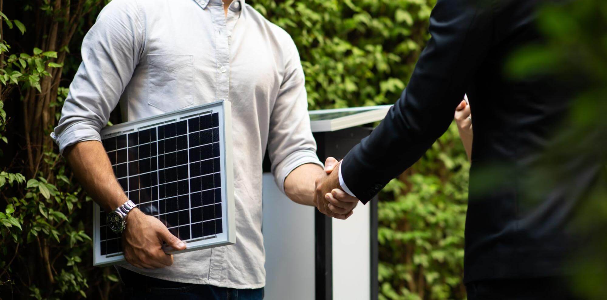 Homem segurando um pequeno painel solar aperta a mão de uma pessoa de terno.