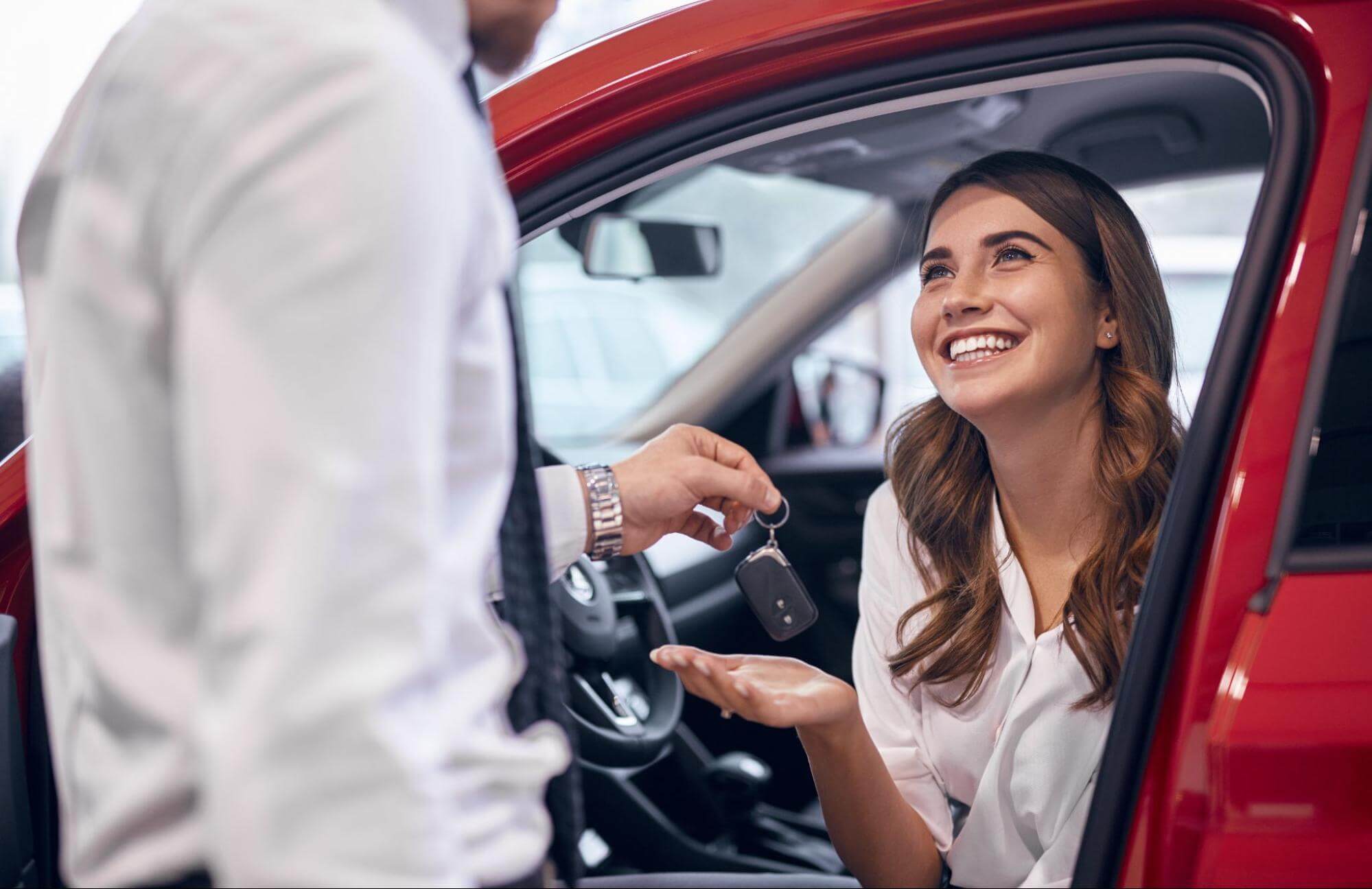 Mulher sentada em veículo vermelho recebe as chaves de automóvel de um um homem à frente. 