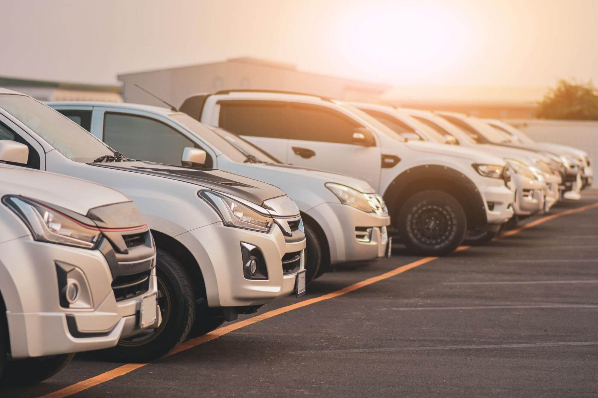Carros enfileirados lado a lado em pátio aberto com pôr-do-sol ao fundo. 