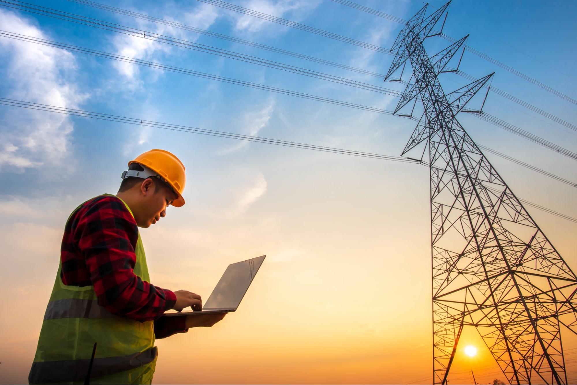 Engenheiro com capacete e laptop trabalhando ao lado de uma torre de energia de alta tensão ao pôr do sol
