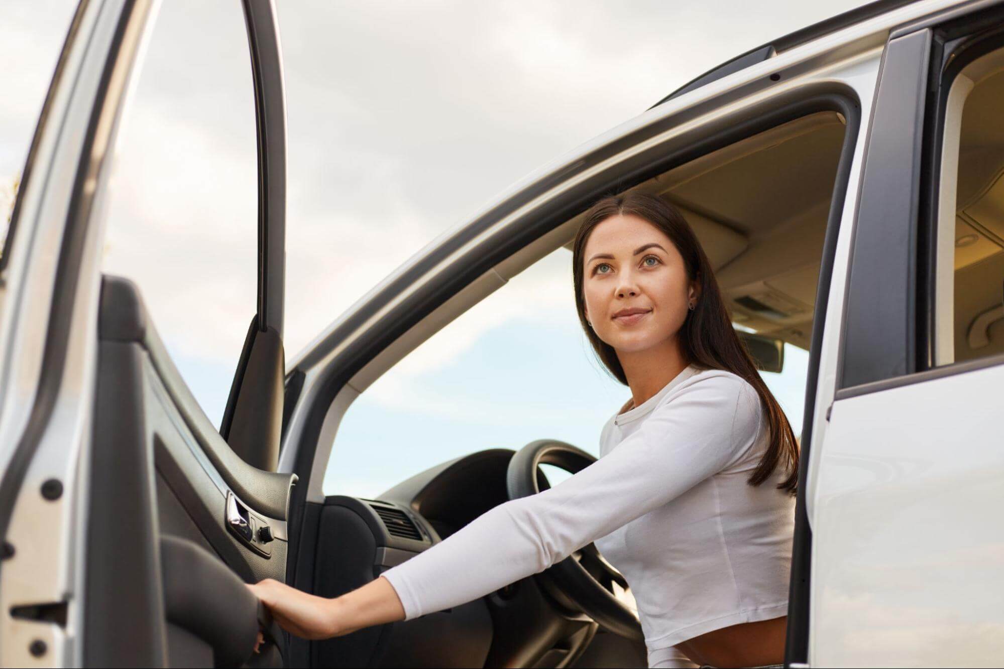 Mulher sentada no banco do motorista de um carro branco com a porta aberta, olhando para cima e para a direita.