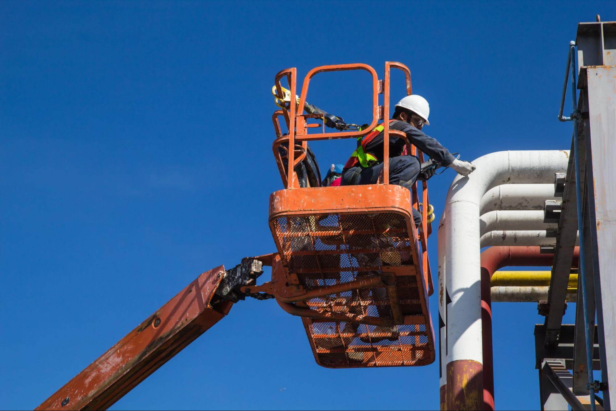 Trabalhador com EPIs trabalhando em plataforma elevatória laranja. 