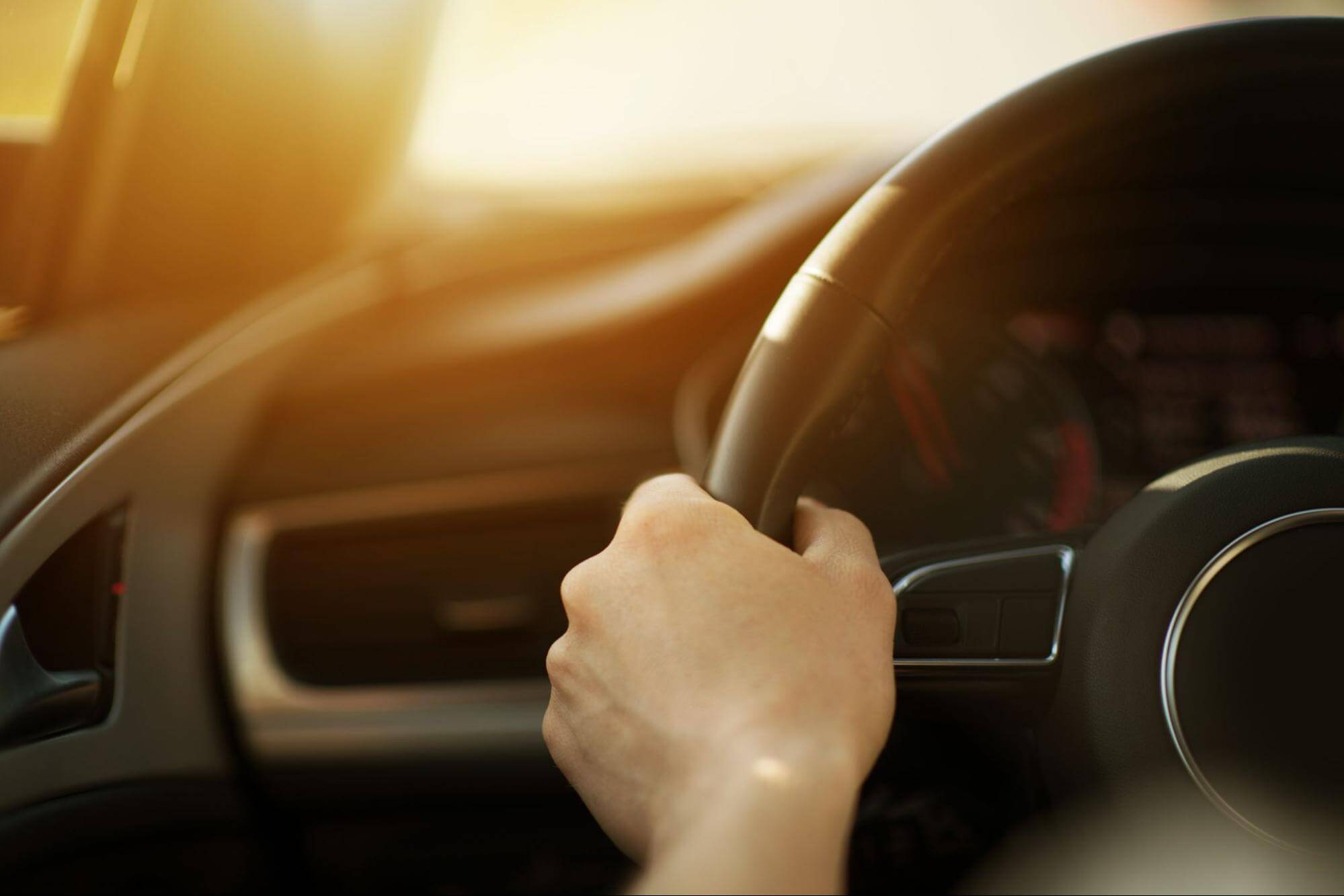 Mão de motorista segurando o volante de um carro, iluminada pela luz do sol ao entardecer.