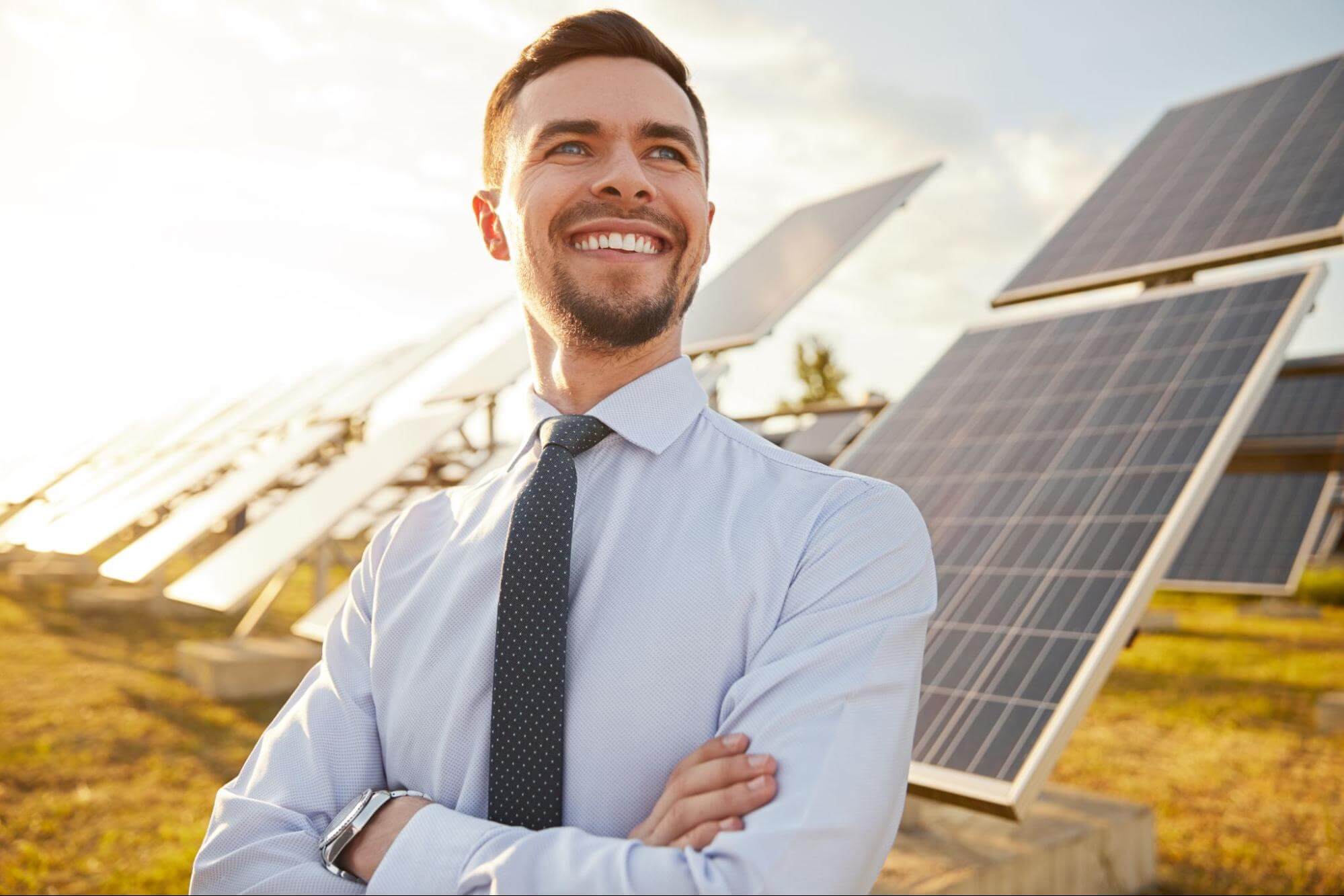 Homem de camisa e gravata sorrindo de braços cruzados em uma usina de energia solar.