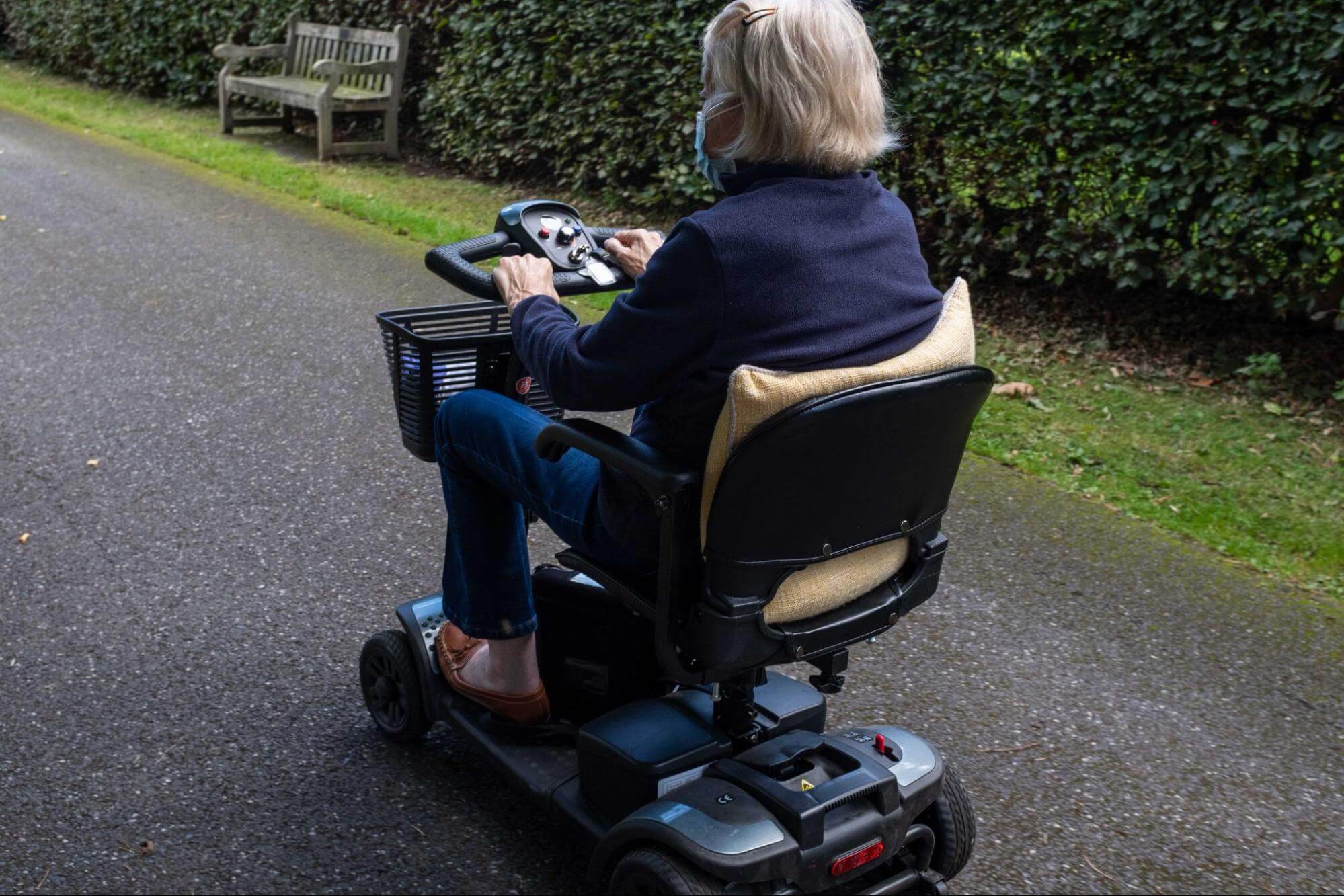 Idosa usando máscara enquanto conduz uma scooter de mobilidade em um caminho ao ar livre.