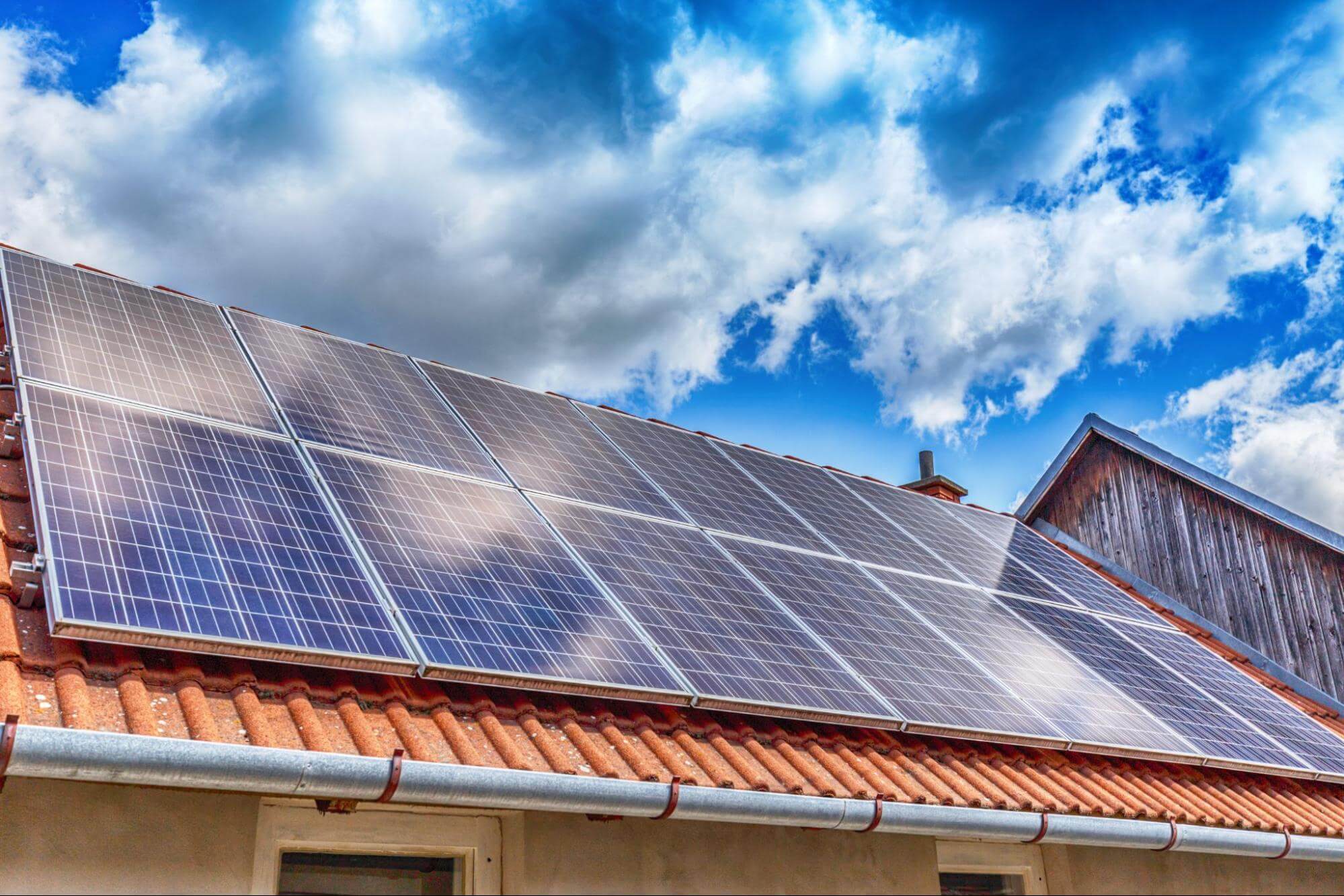 Painéis solares instalados no telhado de cerâmica vermelha de uma casa, sob um céu azul com nuvens brancas.