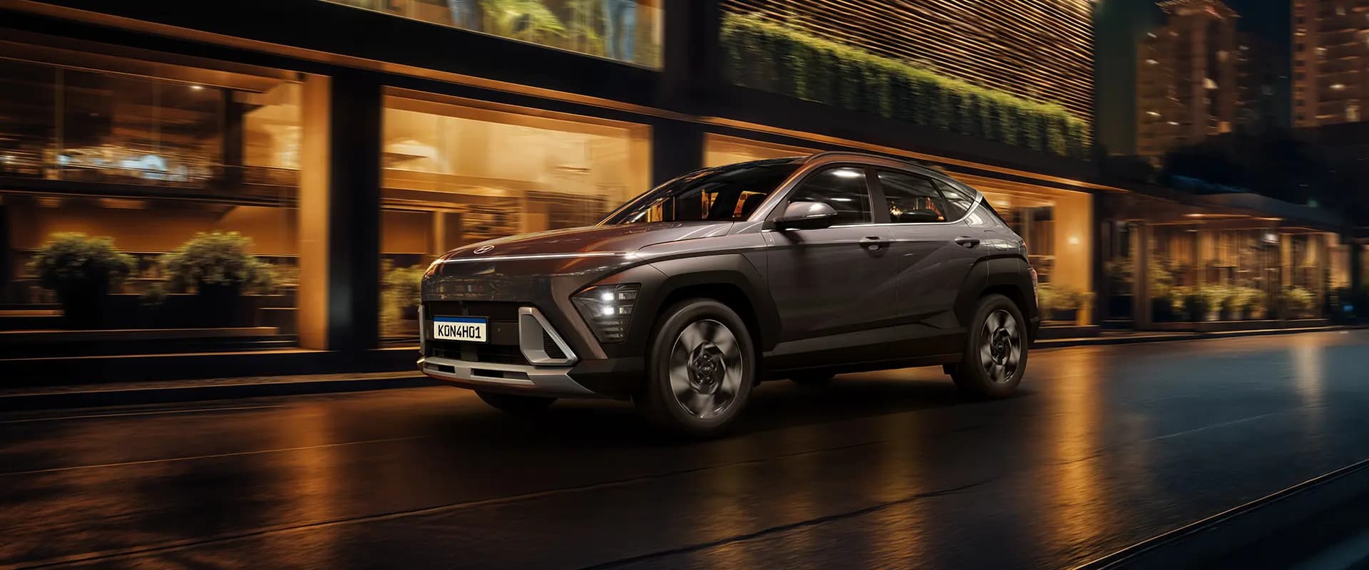 Carro Hyundai Kona EV circulando à noite numa rua urbana iluminada, em frente a edifícios modernos.