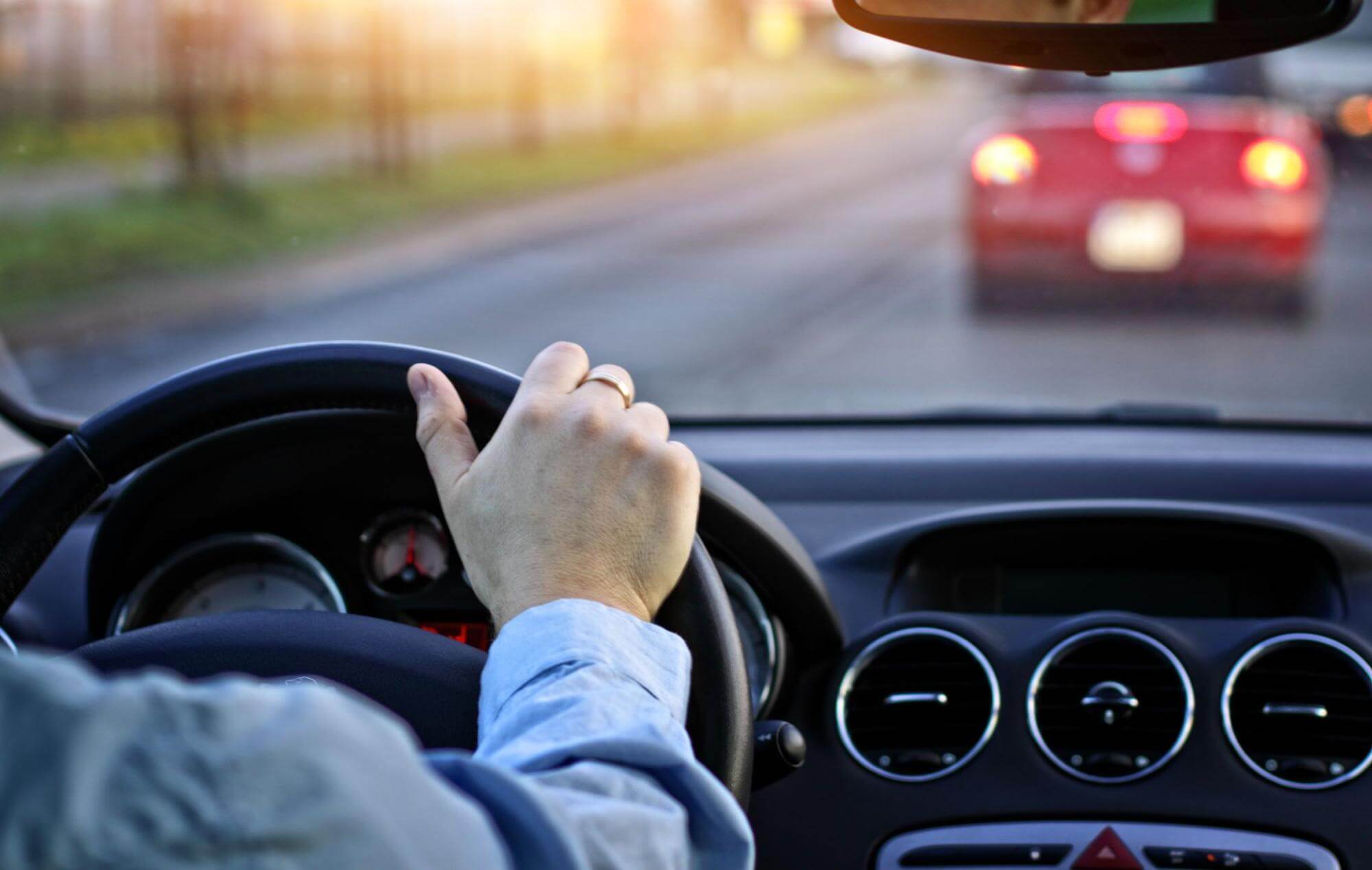 Mão de um motorista segurando o volante de um carro em movimento no trânsito, com outro veículo vermelho à frente.