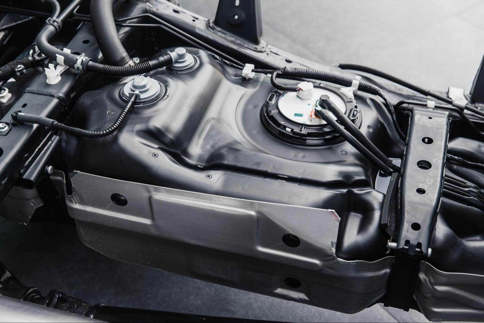 Tanque de combustível metálico montado no chassi de um veículo, mostrando conexões elétricas, mangueiras e tubulações.