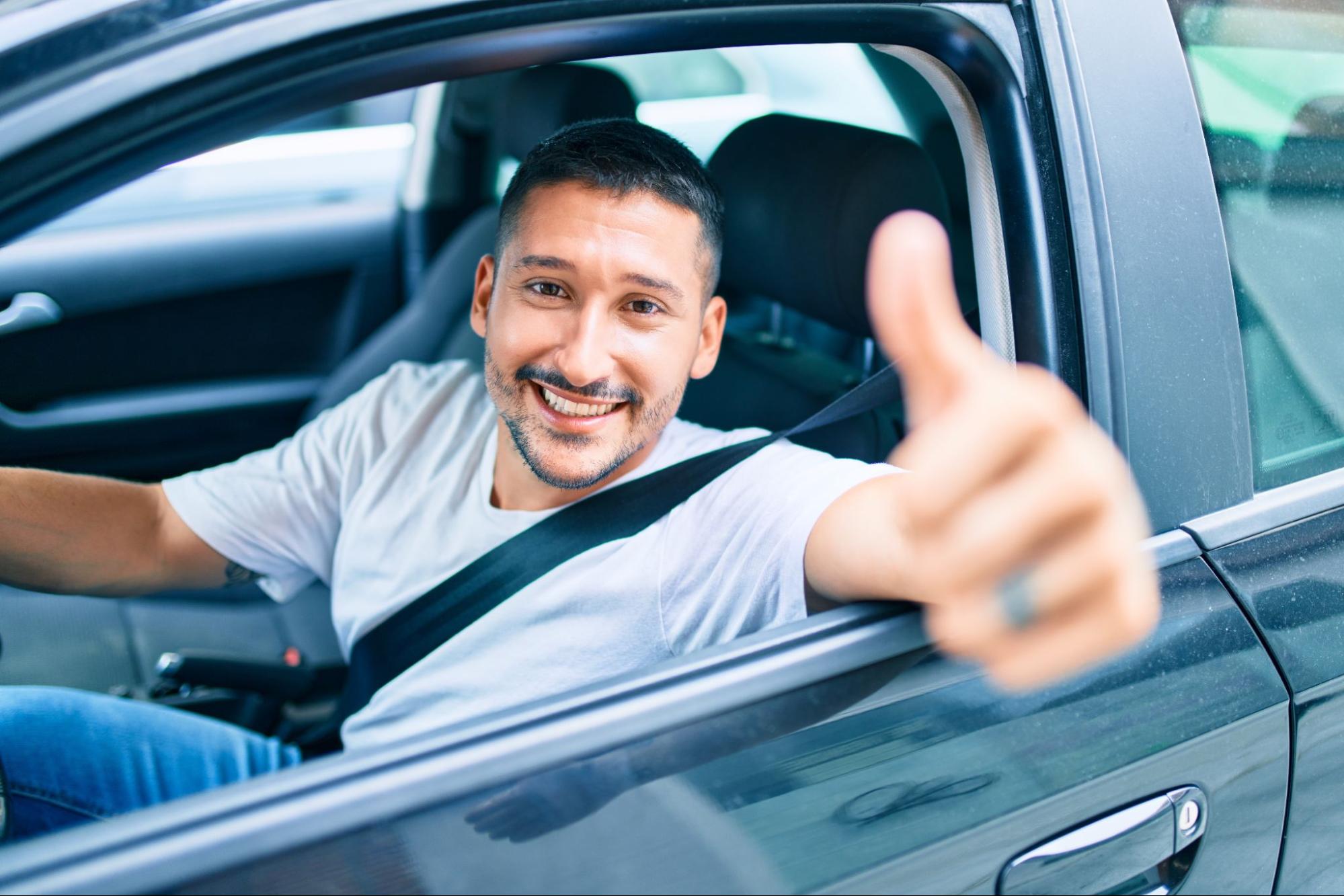 Homem sorrindo dentro do carro e fazendo sinal de positivo pela janela aberta.