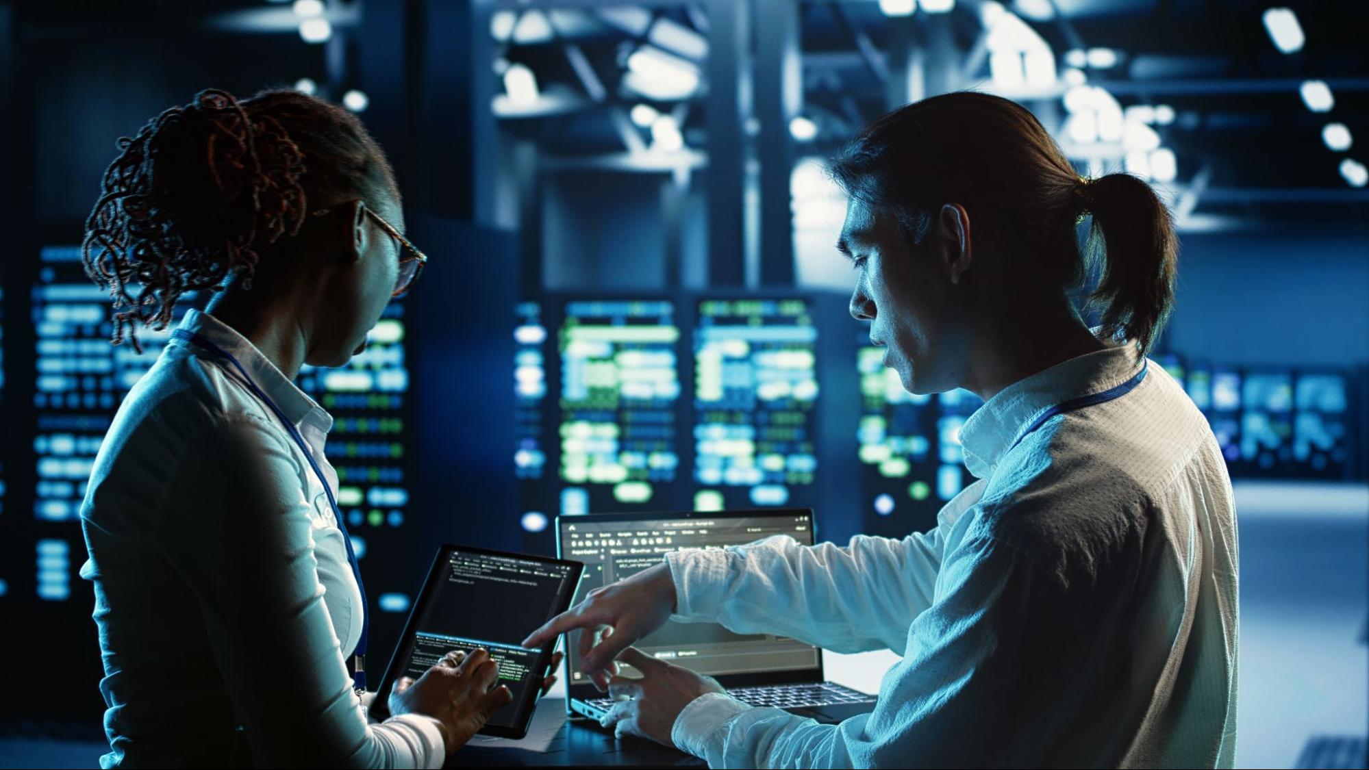 Dois especialistas em TI analisando códigos em um tablet e em um notebook dentro de um data center com iluminação azul.