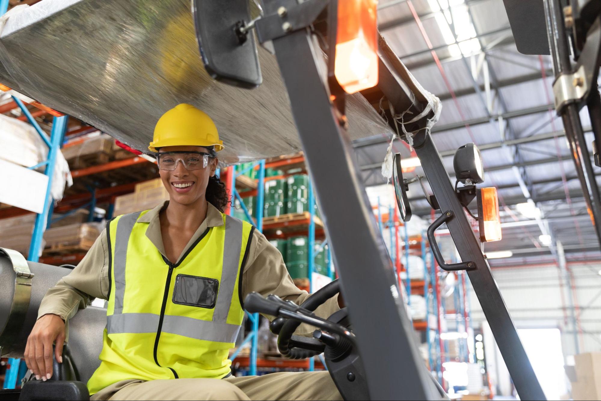Trabalhadora sorridente usando capacete amarelo, óculos de proteção e colete refletor enquanto opera um empilhador num armazém.