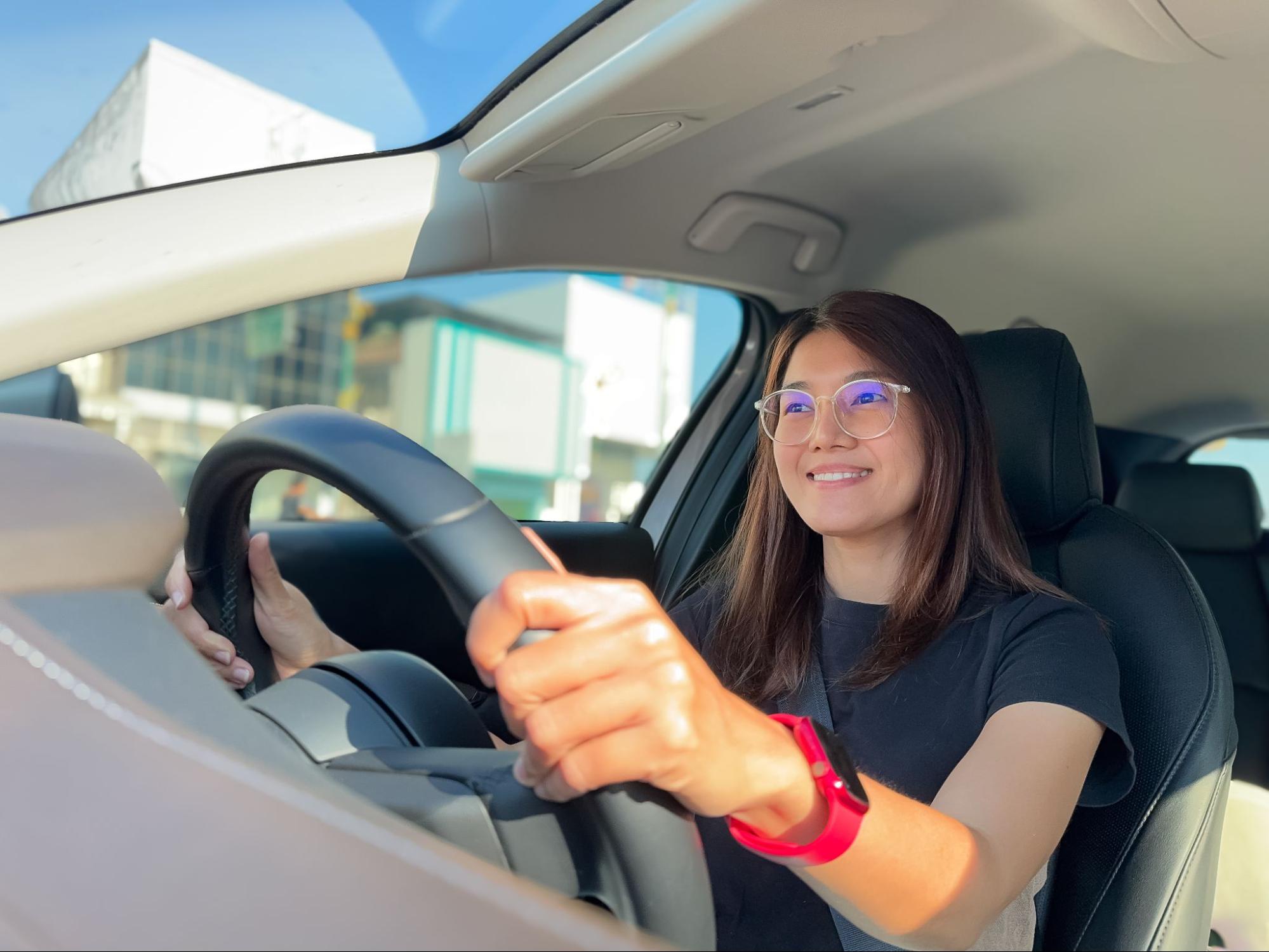 Mulher de óculos sorridente a conduzir um automóvel com as duas mãos no volante.